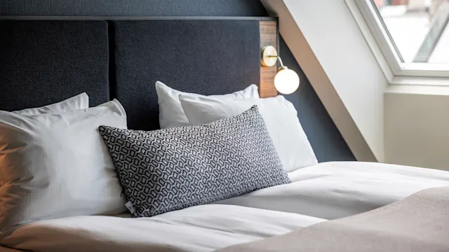 Bed with white sheets and patterned pillow positioned under a modern light fixture, beside a tilted window in a stylish bedroom setting.