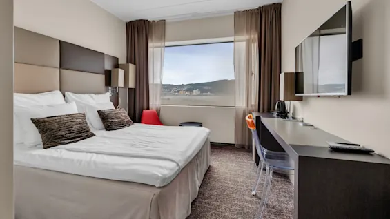 A neatly made bed with brown pillows and bedside lamps sits in a hotel room. A window reveals a city view, and a television hangs on the opposite wall.  