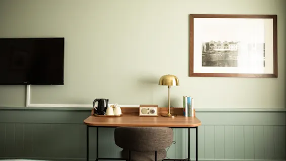 En prydlig skrivbord har en vattenkokare, koppar, en lampa, en vintage radio och böcker. Bredvid hänger en väggmonterad TV och en inramad bild på en ljusgrön vägg.