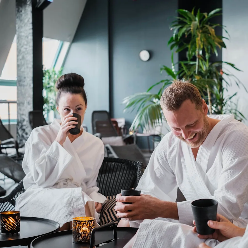 Three people in white robes relax and drink from cups, seated in a spa setting with greenery and ambient lighting around them.