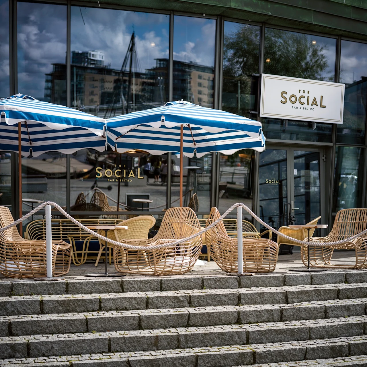 Wickerstolar står under blårandiga parasoller utanför "THE SOCIAL BAR & BISTRO." Stensteg leder till glasdörrar som speglar omgivande byggnader.