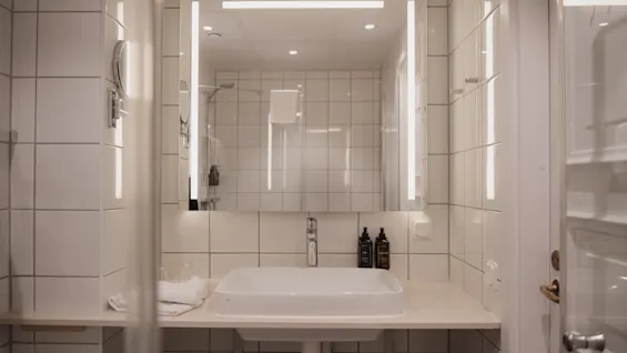 Ett rent och modernt hotellbadrum med en välbelyst spegel ovanför ett vitt handfat, med toalettartiklar och rena handdukar.