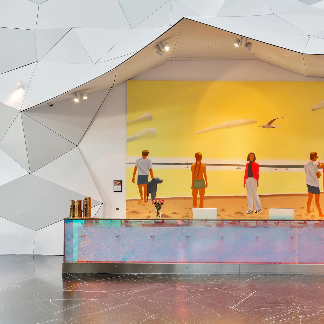 Reception desk adorned with abstract art, facing a mural depicting a beach at sunset. People are on the beach, with modern, angular wall designs surrounding the area. Text is not present.