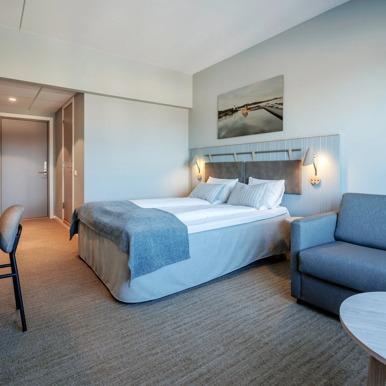 A well-lit hotel room features a neatly made double bed, a gray sofa, a desk with a lamp, and a wall painting, creating a cozy atmosphere.