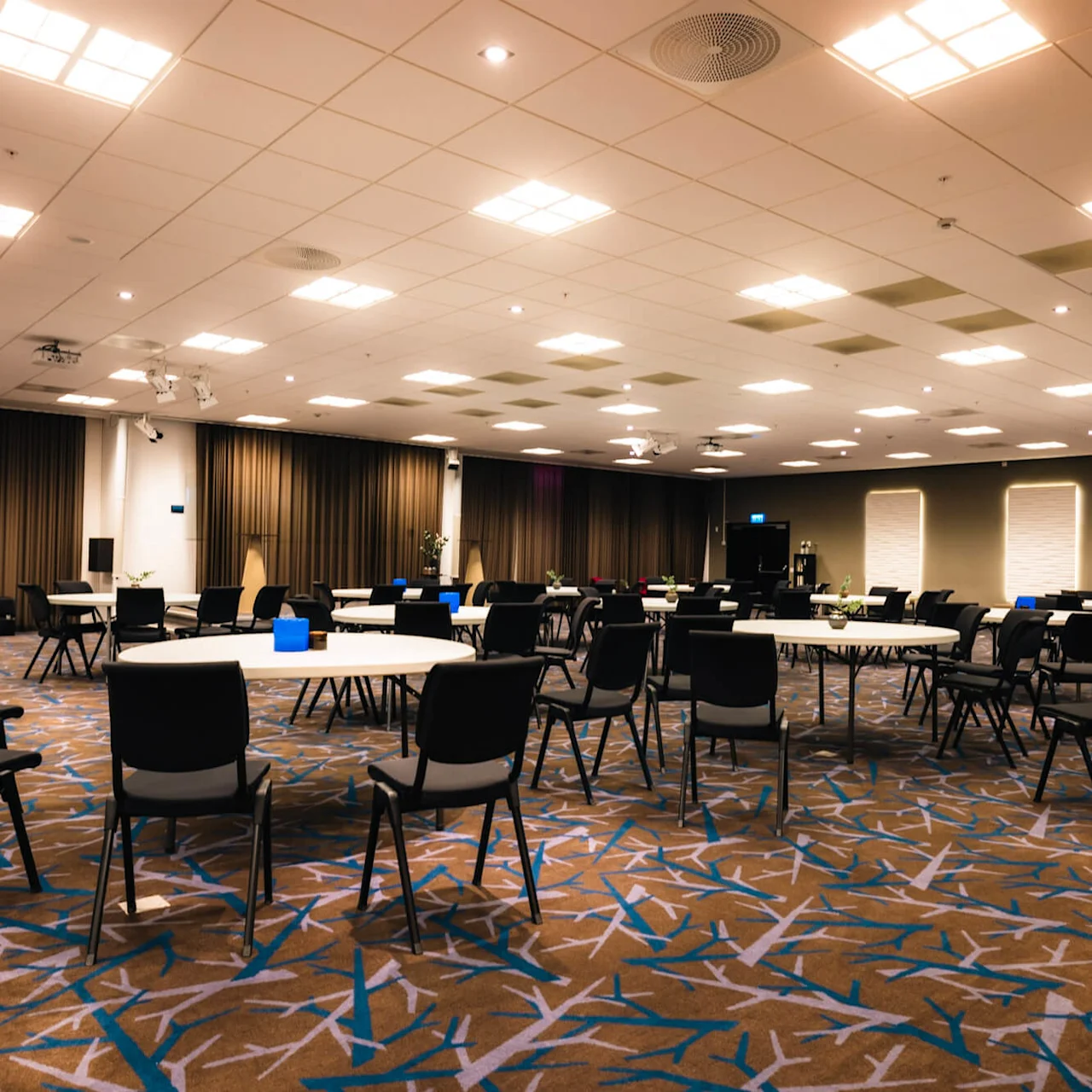 Bord och stolar är arrangerade i ett rymligt konferensrum med mönstrad matta, mjuk belysning och beige gardiner. Taket har flera infällda lampor.