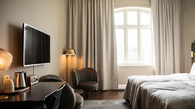 Et hotellrom har en TV på veggen, en koselig seng og et skrivebord med lampe og vannkoker. Gardiner omgir et stort vindu, og skaper en innbydende atmosfære.