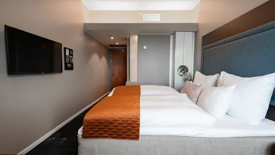 A neatly made double bed with white pillows and orange bed runner in a modern hotel room. A flat-screen TV is mounted on the wall; hallway and door are visible.