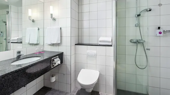 A bathroom features a wall-mounted sink with a granite countertop, a glass-enclosed shower, and a wall-hung toilet. White tiles and towels provide a clean, modern appearance.