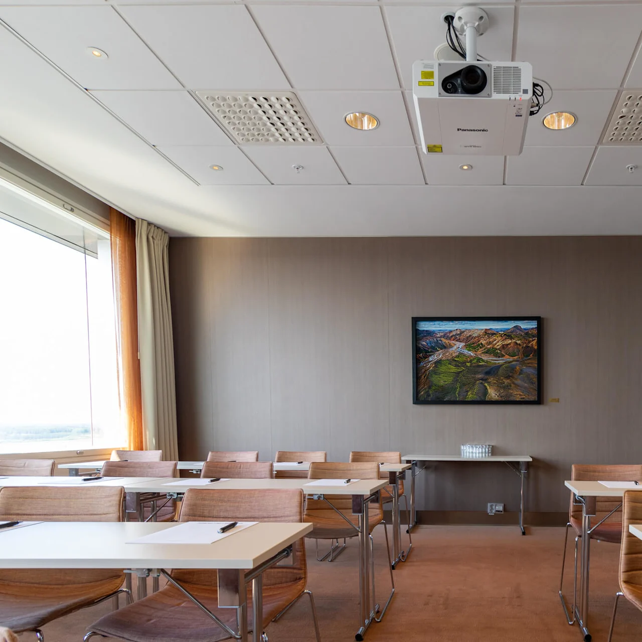 Classroom setting with neat rows of tables and chairs, projector ceiling-mounted. Mountain landscape painting on wall, coatracks to the right, door clock. Papers and pens on tables.