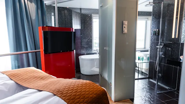 Bed with orange quilt facing a red TV stand in a modern hotel room, beside a bathroom with glass walls and a freestanding bathtub.