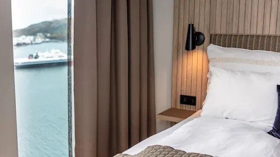 A bed with white linens and a textured blanket is against a wooden headboard in a room with curtains. Outside, a ferry is visible on the water.