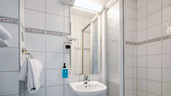 Sink mounted on a tiled wall with a mirror above, adjacent to a shower enclosure. Nearby, a towel hangs on a ring. A dispenser reads "HAND WASH".