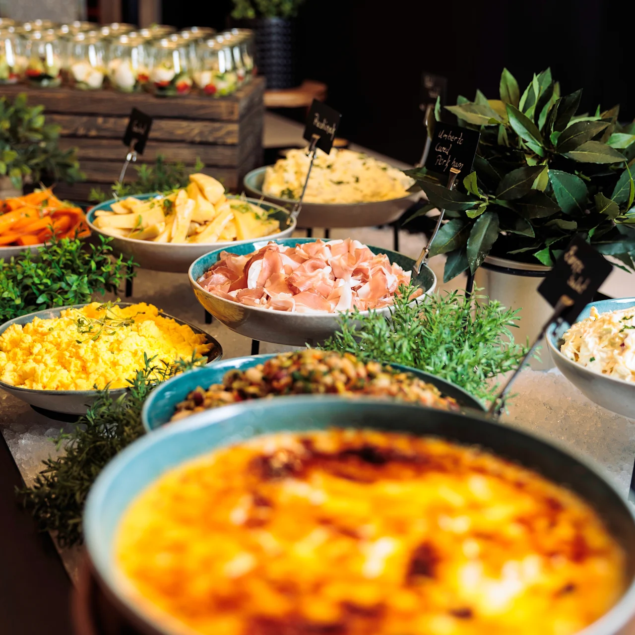 Bowls of assorted foods, including scrambled eggs and sliced meats, are arranged on a buffet table, surrounded by greenery. Small signs label the dishes. Background includes jars of desserts.