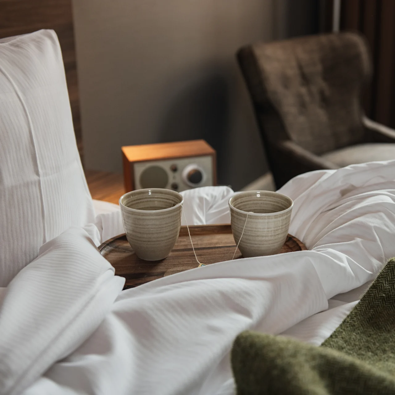 Ceramic cups sit on a wooden tray atop a rumpled, white bed. Nearby, a brown chair and a small wooden speaker add to the cozy atmosphere.
