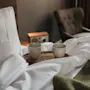 Ceramic cups sit on a wooden tray atop a rumpled, white bed. Nearby, a brown chair and a small wooden speaker add to the cozy atmosphere.