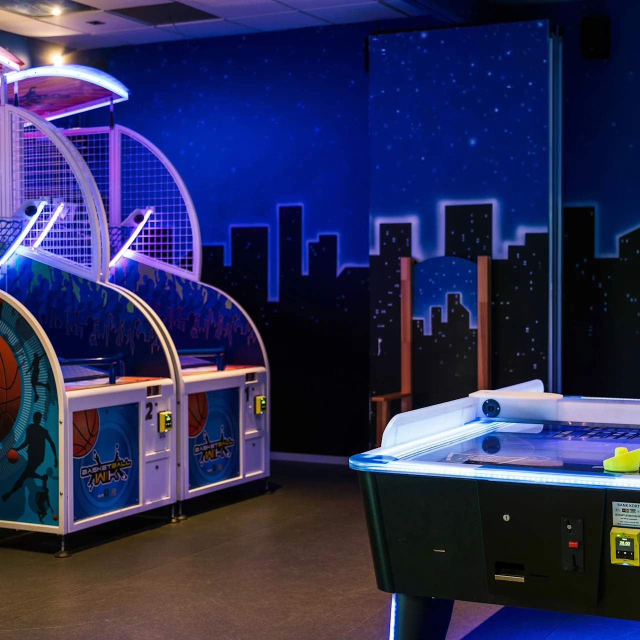 Arcade basketball hoops emit neon lights; a nearby air hockey table has an illuminated edge. The backdrop is a starry sky and city skyline. 