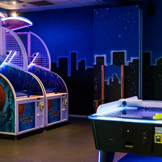 Arcade basketballkurver avgir neonlys; et nærliggende airhockeybord har en opplyst kant. Bakgrunnen er en stjerneklar himmel og bysilhuett.