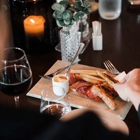 En person nyter et forrettsfat med spekemat, ost og brød, akkompagnert av vin i en koselig, levende lyssetting.