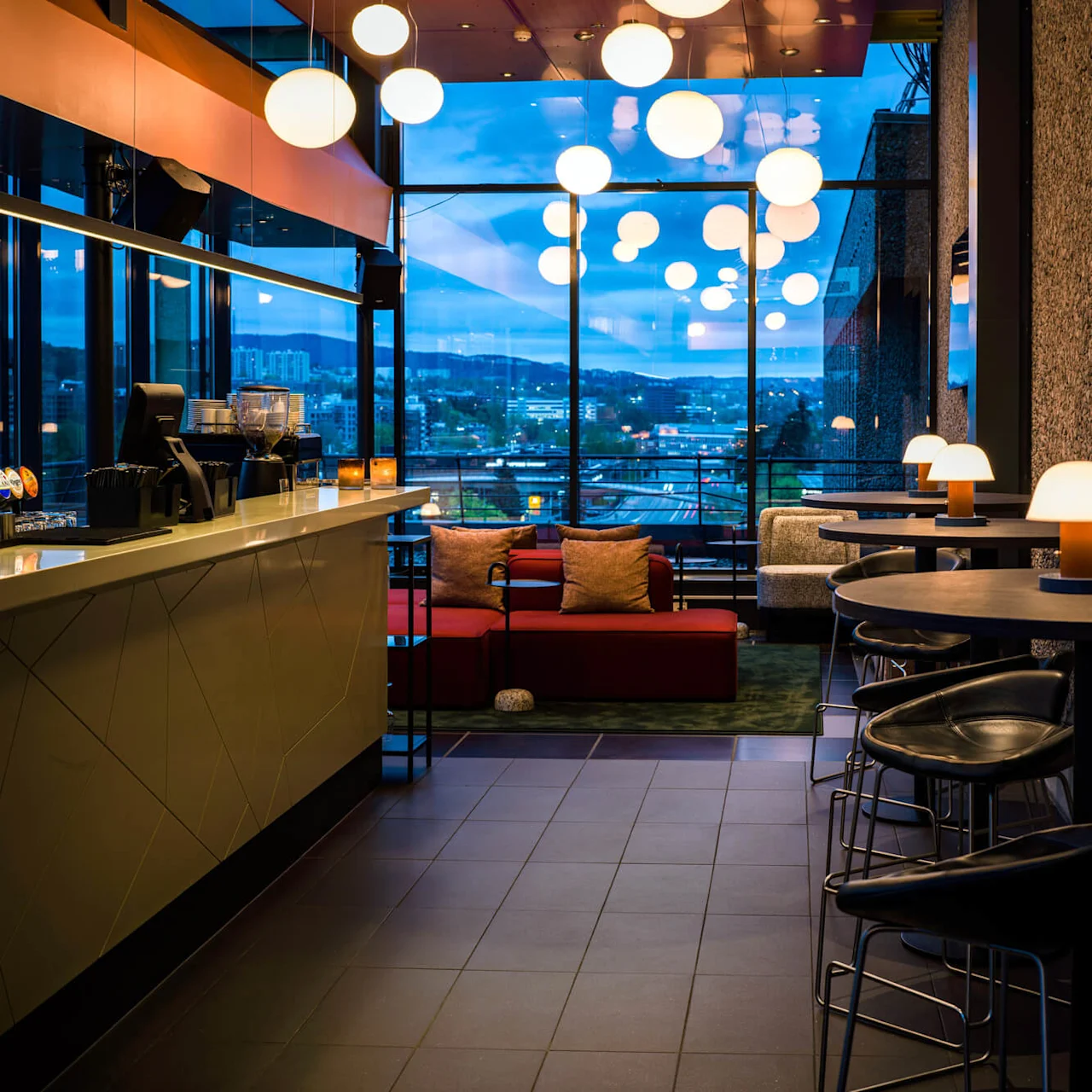 A modern bar with hanging spherical lights, features a sleek counter on the left, cushioned seating in the center, and views of a city skyline through large windows.