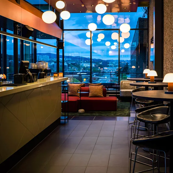 En moderne bar med hengende sfæriske lys, har en elegant disk til venstre, polstret sitteplasser i midten og utsikt over byens skyline gjennom store vinduer.