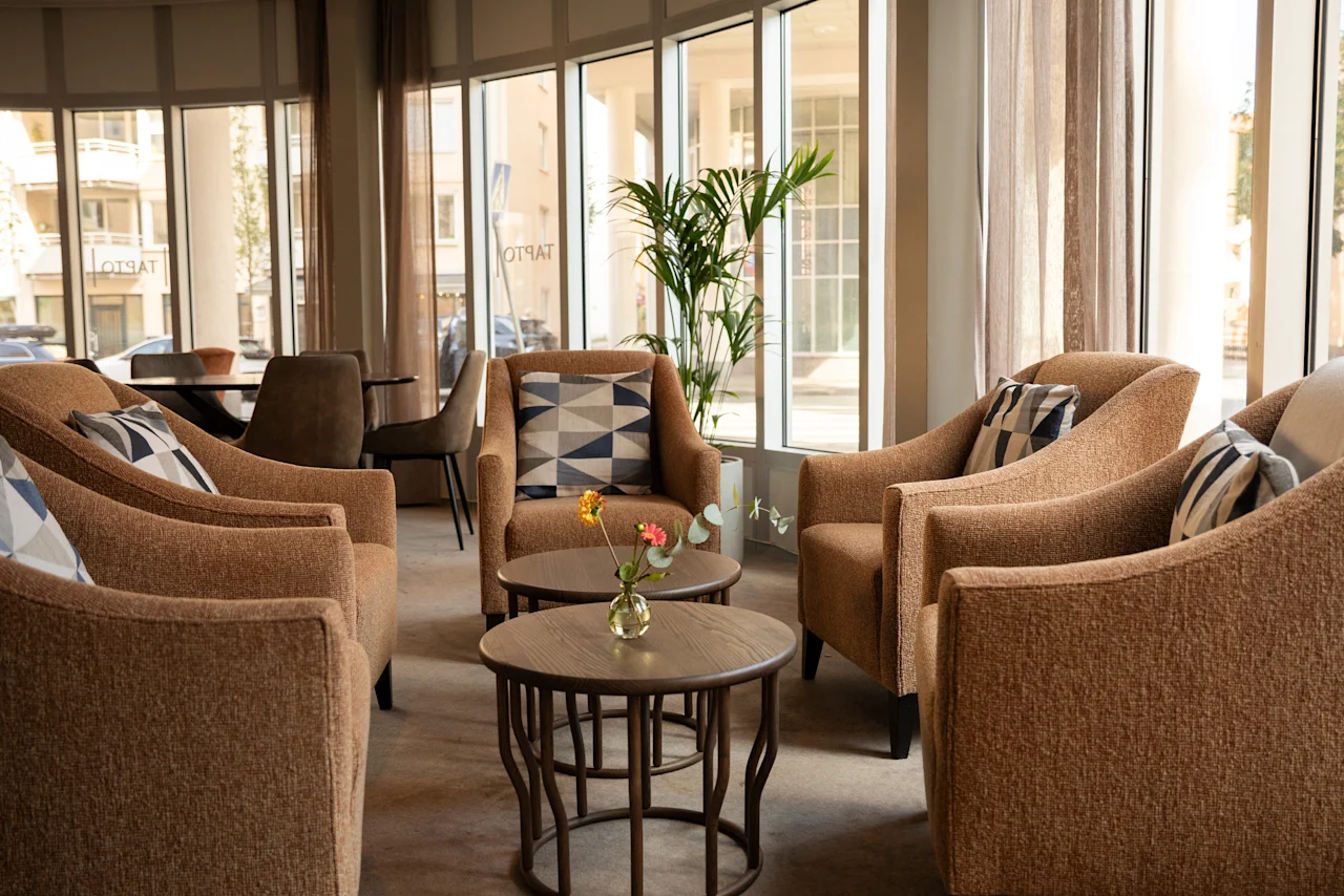 En mysig hotellounge med bekväma fåtöljer och små bord, badad i naturligt ljus från stora fönster.