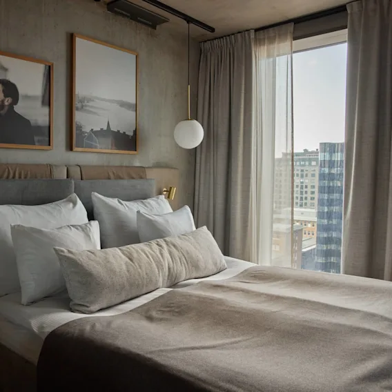 A neatly made bed with gray and white bedding sits in a modern room. Framed photographs hang above, with city buildings visible through the large window.