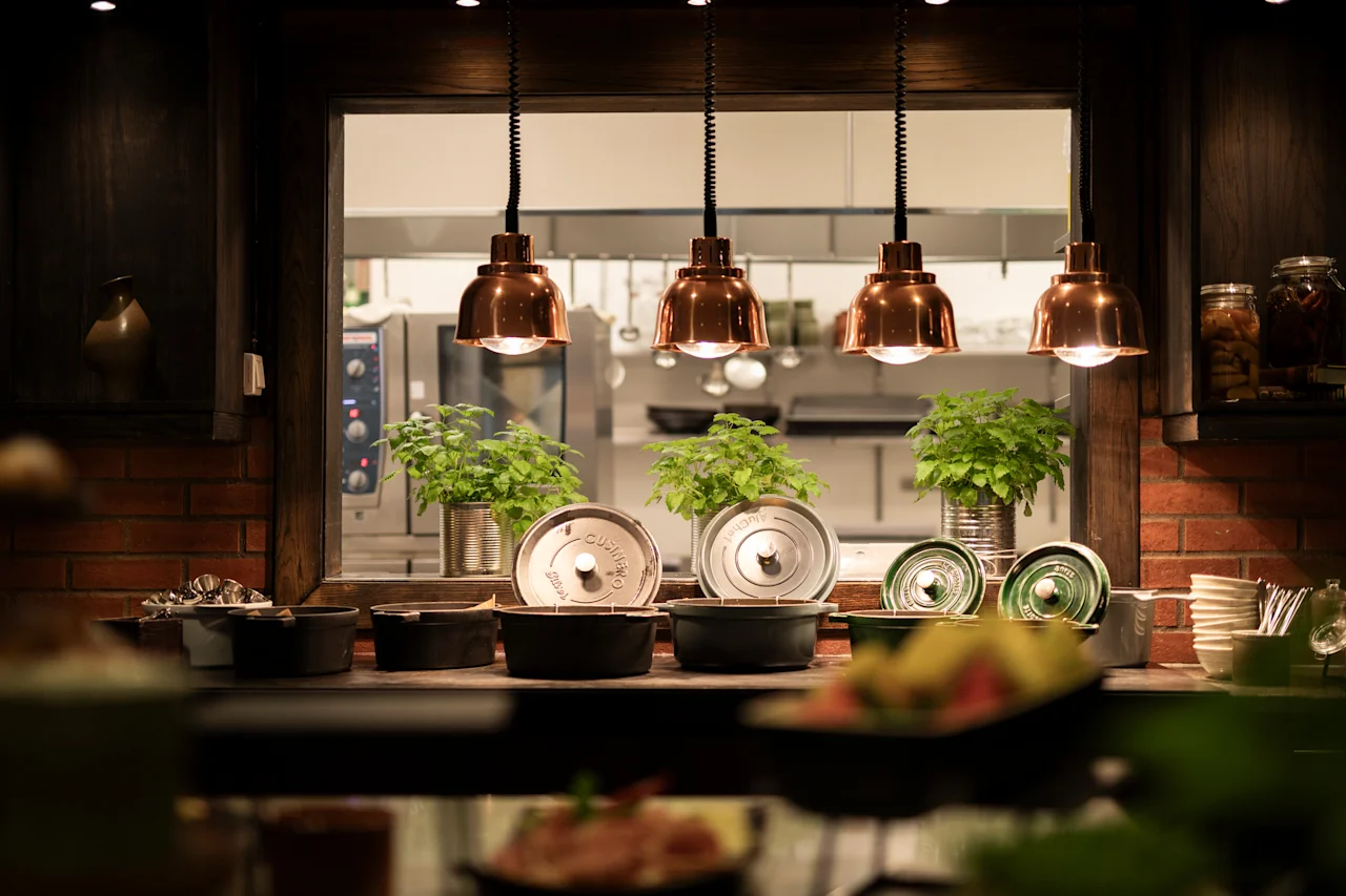 A warm and inviting food service area with a glimpse into a busy kitchen, featuring copper lamps and fresh plants.