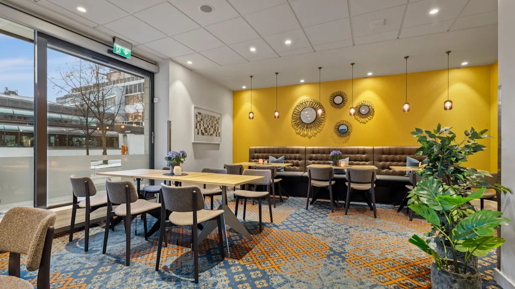 Ett färgglatt och inbjudande mat- eller loungeområde med gula väggar, mönstrad matta och gott om sittplatser. En utgångsskylt är synlig.