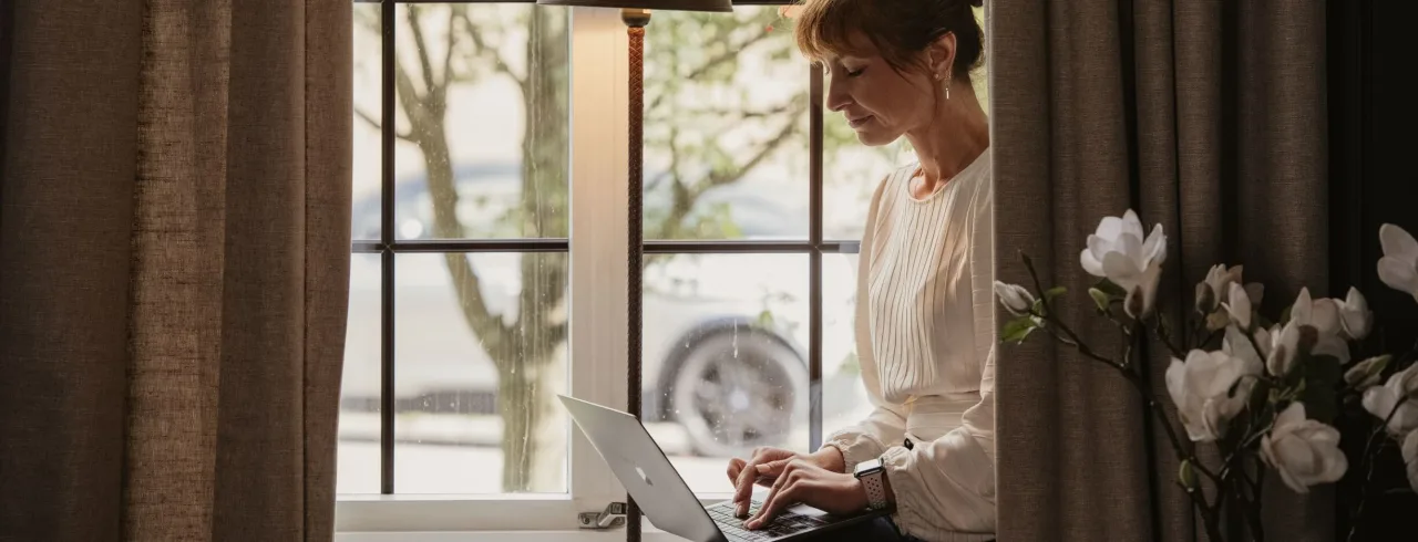 A woman works on a laptop by a window, with soft lighting and curtains framing the scene.