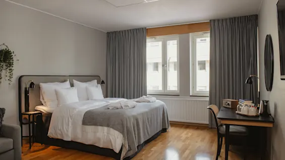 En pent, oppredd seng med hvitt sengetøy og grått teppe er i et romslig, moderne hotellrom. Lys strømmer gjennom store vinduer, ved siden av et skrivebord og stol.