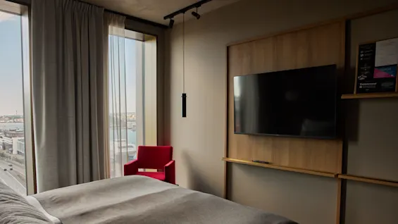 En stor TV monterad på en träpanel finns i ett modernt hotellrum. En grå sängöverkast täcker sängen och en röd fåtölj står nära fönstret med utsikt över stadsmiljön.
