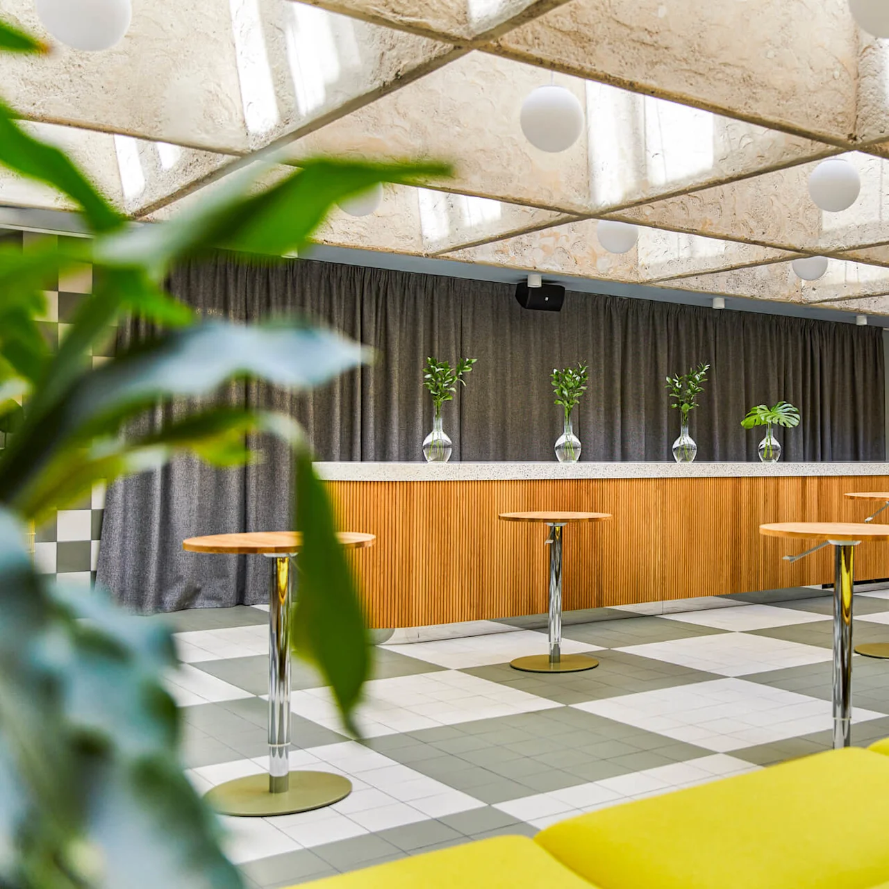 Et bardiskområde med høye, runde borde og en disk dekoreret med planter i glasvaser. Indretningen inkluderer ternet gulv, et reflekterende loft og grå gardiner.