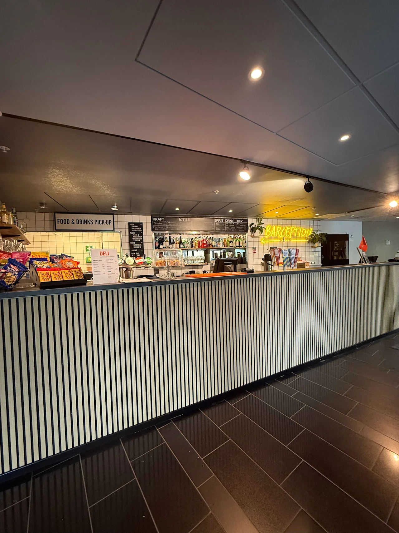En modern bar- och receptionsdisk med snacks, drycker och skyltar för mat- och dryckesupphämtning samt delitjänster.