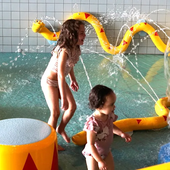 Barn leker glatt i en grund pool med vatten som stänker från färgglada lekstrukturer.