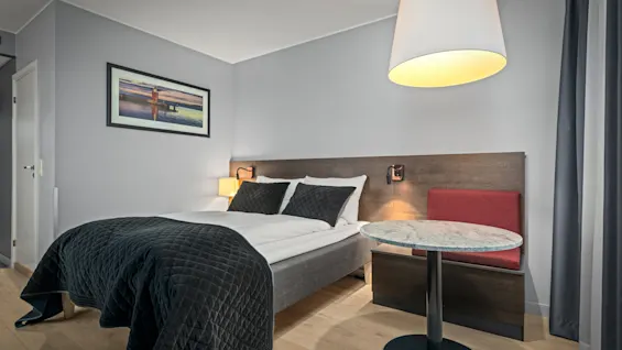 En pænt redt seng med et mørkt tæppe står mod et træhovedgærde, ledsaget af et lille rundt bord og lampe i et moderne hotelværelse.