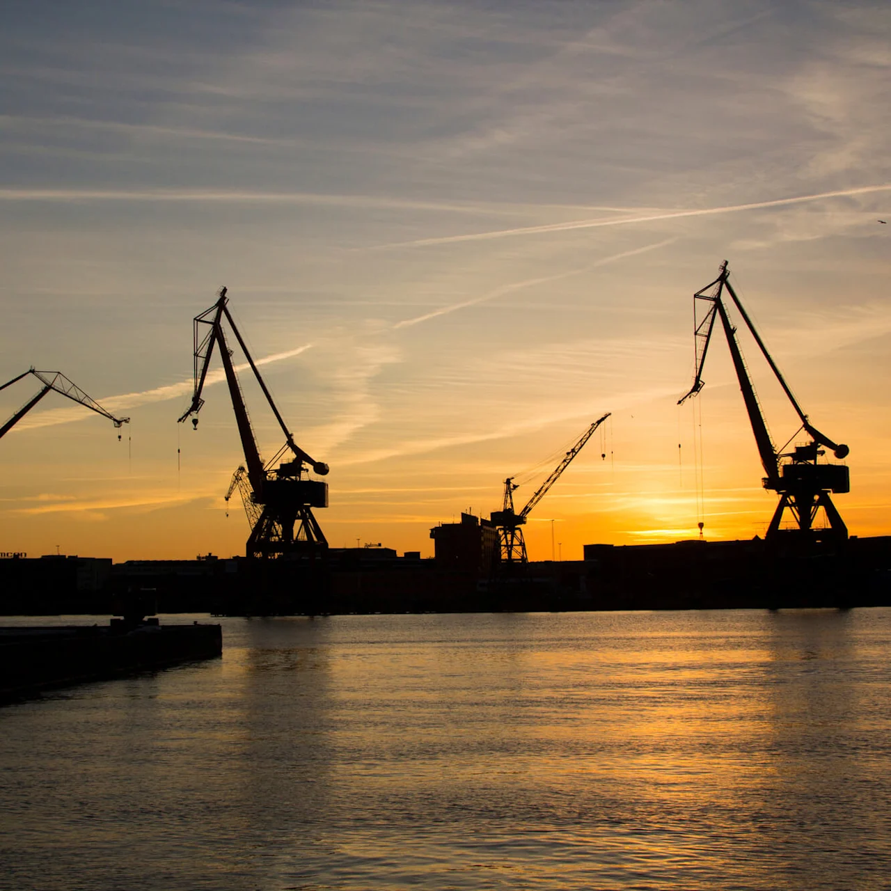 Cranes silhouetted against a sunset, stretching over a calm waterfront in an industrial harbor setting.