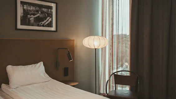 En säng är prydligt bäddad bredvid en modern golvlampa, under ett inramat svartvitt foto. En stol och ett stort fönster med fördragna gardiner kompletterar denna mysiga hotellrumsmiljö.