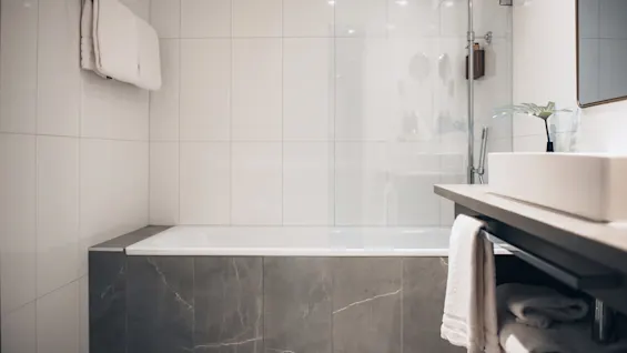 A white bathtub is situated against a tiled wall, surrounded by a minimalist bathroom with a towel rack, a countertop sink, and a small plant in a vase.