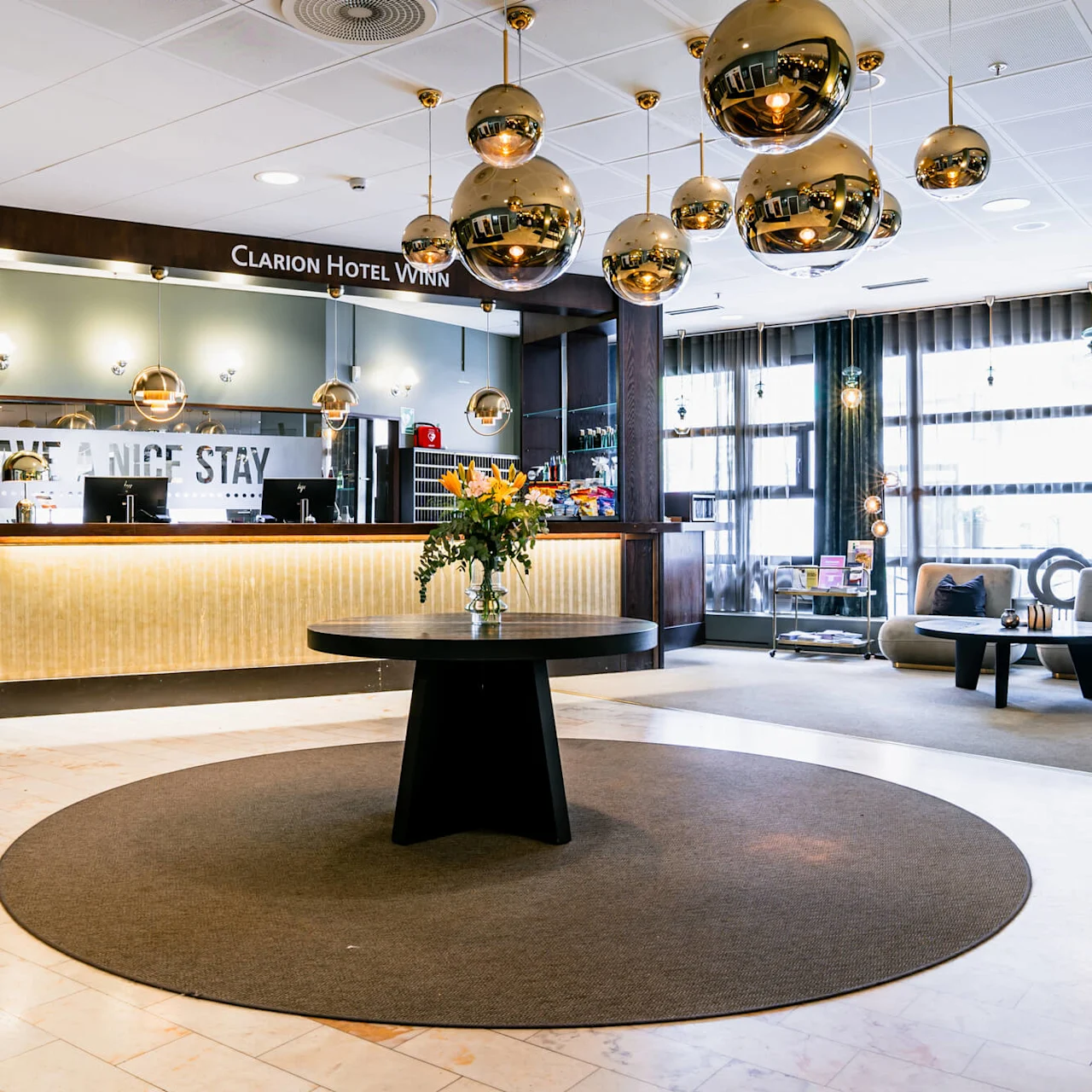 En hotellobby med et sort rundt bord og blomster, omgivet af moderne siddepladser og gyldne sfæriske lamper. Skilte siger "Clarion Hotel Winn," "EXPRESS CHECK OUT," og "HAVE A NICE STAY."