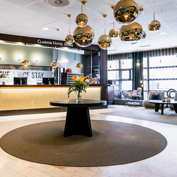 Ett hotellobby med ett svart runt bord och blommor, omgiven av moderna sittplatser och gyllene sfäriska lampor. Skyltar lyder "Clarion Hotel Winn," "EXPRESS CHECK OUT," och "HAVE A NICE STAY."