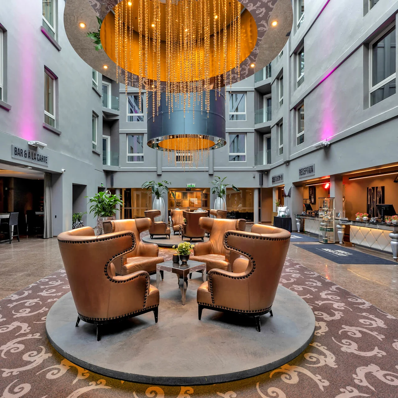 En cirkel af brune læderstole omgiver et lille bord under en stor lysekrone i en hotellobby med skilte, hvor der står "BAR À LA CARTE" og "RECEPTION".