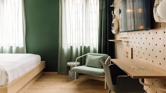 A modern hotel room with a bed, green armchair, wooden desk, and a pegboard wall with a TV. 