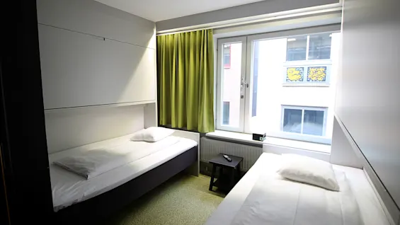 Valoisa hotellihuone, jossa on kaksi yhden hengen sänkyä, vihreät verhot ja ikkuna.