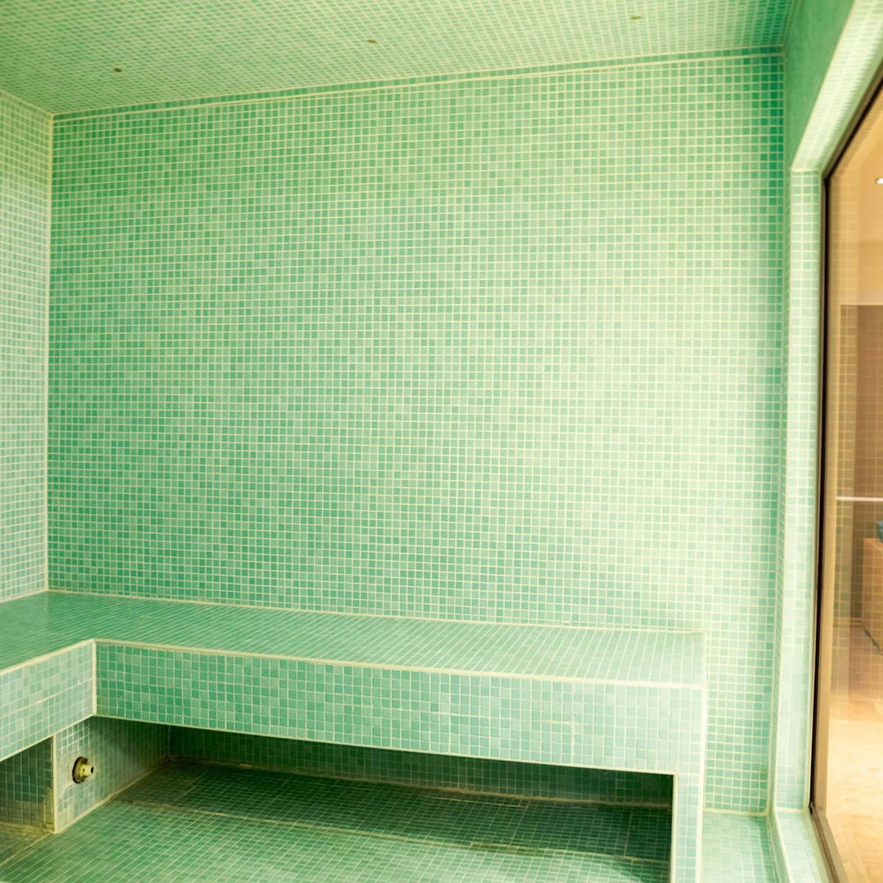 Green-tiled steam room with a bench lines the left wall; a transparent partition reveals an adjoining area with warm lighting.