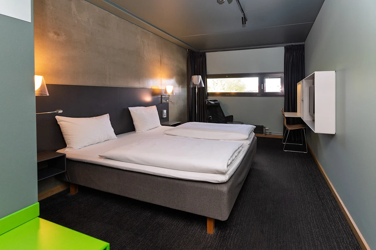 En prydligt bäddad dubbelsäng med vita sängkläder står i ett modernt hotellrum med mörka väggar och ett litet skrivbord nära ett fönster, belyst av väggmonterade lampor.