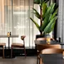 Et koselig restaurantinteriør med mørke bord, brune stoler og en stor grønn plante. Myk belysning fra stearinlys skaper en varm atmosfære.
