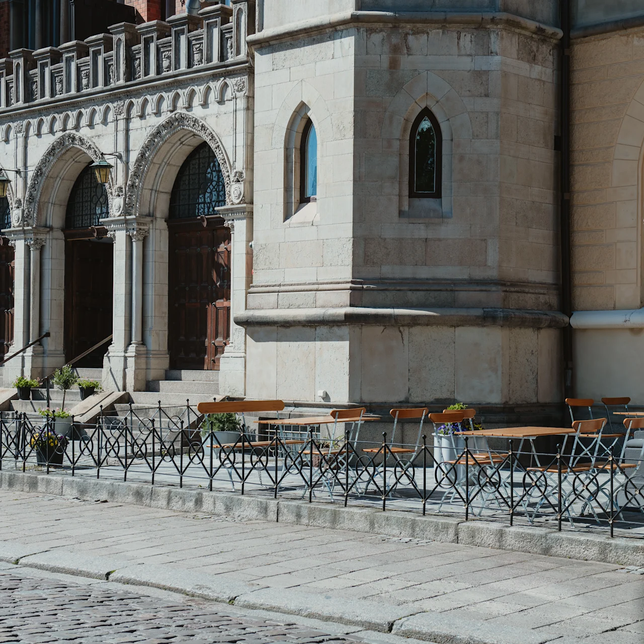 Utomhusbord och stolar från en kafé är prydligt arrangerade längs en trottoar, intill en historisk stenbyggnad med bågformade dörröppningar. Krukväxter och järnräcken bidrar till den inbjudande atmosfären.
