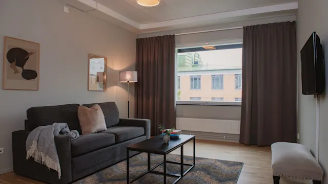 Grå soffa med kuddar och filt, placerad i ett modernt vardagsrum. Ett soffbord står framför, och ett stort fönster med gardiner visar en byggnad utanför.