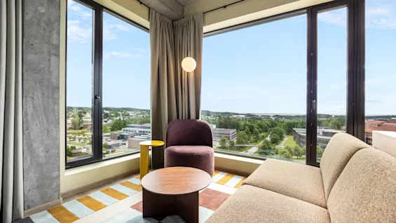 A bright, modern living space with large windows overlooking a green landscape and city, featuring a sofa, armchair, and coffee table.