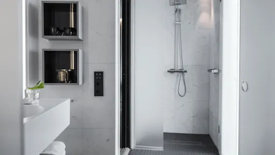 Et moderne bad med en glasbruser og regnbruserhoved. Væghylder indeholder toiletartikler, og en håndvask med en plante er synlig, omgivet af hvide fliser.